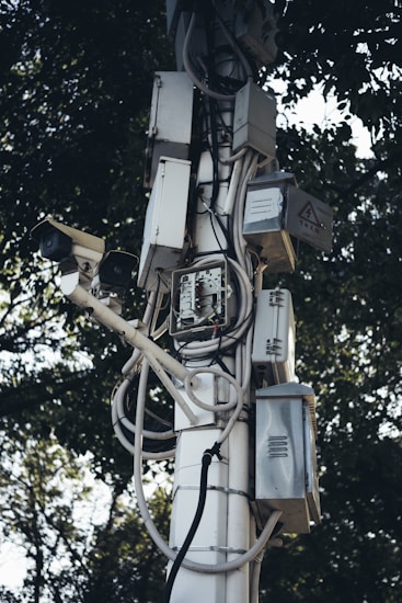 A tall pole is covered with multiple electrical boxes and surveillance cameras, intertwined with various wires and cables. The equipment is mounted in an organized manner, with trees in the background providing a natural contrast.