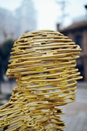 A sculpture composed of yellow metal wires intricately woven to form a human head. The wires create a hollow structure with a mesh-like appearance, set against an urban backdrop with buildings and blurry skies. The design exhibits a blend of abstract art and human form.