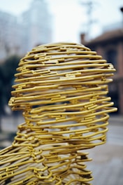 A sculpture composed of yellow metal wires intricately woven to form a human head. The wires create a hollow structure with a mesh-like appearance, set against an urban backdrop with buildings and blurry skies. The design exhibits a blend of abstract art and human form.