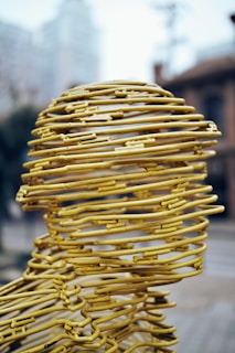 A sculpture composed of yellow metal wires intricately woven to form a human head. The wires create a hollow structure with a mesh-like appearance, set against an urban backdrop with buildings and blurry skies. The design exhibits a blend of abstract art and human form.
