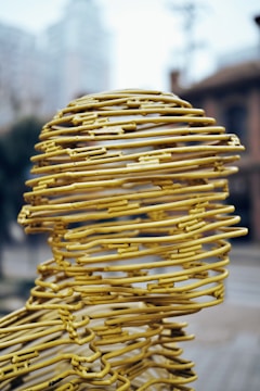 A sculpture composed of yellow metal wires intricately woven to form a human head. The wires create a hollow structure with a mesh-like appearance, set against an urban backdrop with buildings and blurry skies. The design exhibits a blend of abstract art and human form.