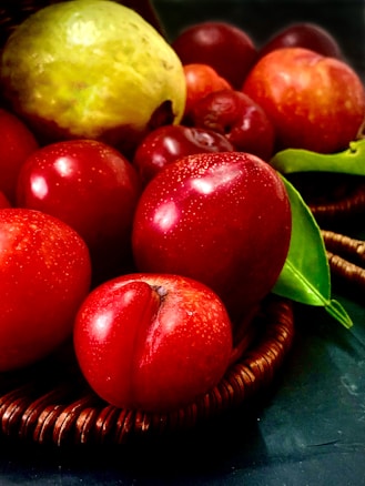 A basket filled with ripe and vibrant fruits, predominantly red apples, a pear, and other similar fruits. The textures and colors are vivid, capturing the freshness and succulence of the produce. A leafy green element adds contrast to the red tones.