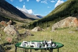A scenic mountain valley with a vibrant green meadow scattered with large rocks. In the foreground, a portable table holds a chessboard with pieces arranged for a game.