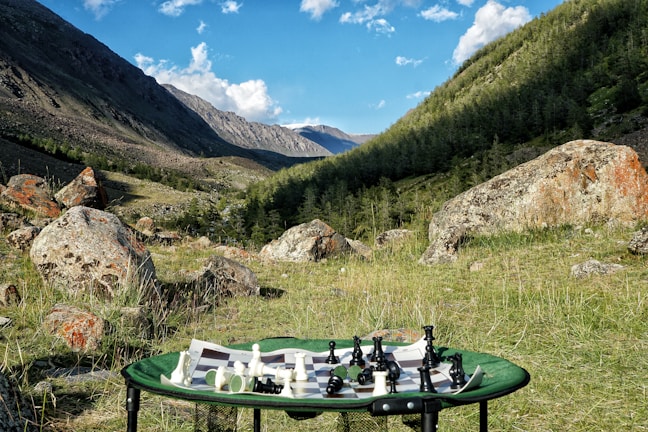 A scenic mountain valley with a vibrant green meadow scattered with large rocks. In the foreground, a portable table holds a chessboard with pieces arranged for a game.