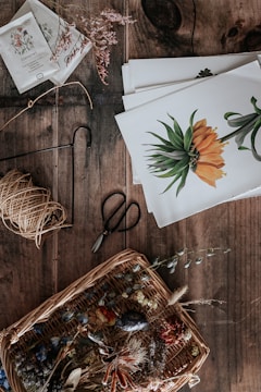 A rustic wooden table holds a collection of botanical and crafting materials. There's a wicker basket filled with various dried flowers and leaves, creating a natural and earthy arrangement. Next to the basket, a pair of vintage scissors rests alongside some twine. Several botanical illustrations featuring vibrant flowers are neatly stacked, providing a theme of botanical art and crafts. Seed packets and small sprigs of dried plants are scattered, hinting at a gardening or artistic project.