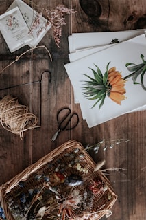 A rustic wooden table holds a collection of botanical and crafting materials. There's a wicker basket filled with various dried flowers and leaves, creating a natural and earthy arrangement. Next to the basket, a pair of vintage scissors rests alongside some twine. Several botanical illustrations featuring vibrant flowers are neatly stacked, providing a theme of botanical art and crafts. Seed packets and small sprigs of dried plants are scattered, hinting at a gardening or artistic project.