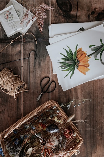 A rustic wooden table holds a collection of botanical and crafting materials. There's a wicker basket filled with various dried flowers and leaves, creating a natural and earthy arrangement. Next to the basket, a pair of vintage scissors rests alongside some twine. Several botanical illustrations featuring vibrant flowers are neatly stacked, providing a theme of botanical art and crafts. Seed packets and small sprigs of dried plants are scattered, hinting at a gardening or artistic project.