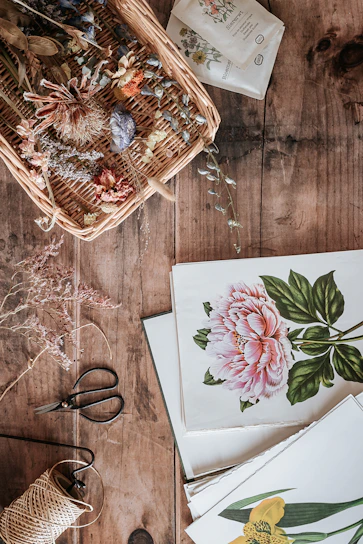 A wooden table adorned with botanical elements, including a wicker basket filled with dried flowers and a selection of botanical prints. Next to the basket is a pair of black garden scissors, dried plant stems, and a roll of string. The prints showcase detailed floral illustrations with vibrant colors.
