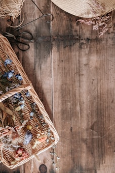 A rustic wooden table features a wicker basket filled with an assortment of dried flowers, including blue and red blooms. Nearby, a coil of twine and a pair of black metal scissors are placed, suggesting a crafting or floral arrangement activity. A woven straw hat with dried floral decorations is partially visible, adding a natural and artisanal feel to the scene.