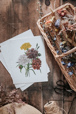 A wicker basket filled with an assortment of dried flowers sits on a rustic wooden surface. Next to the basket, there are sheets of paper with botanical illustrations, showcasing detailed drawings of colorful flowers. A spool of twine, scissors, and a straw hat are also visible, adding to the vintage and handcrafted aesthetic.
