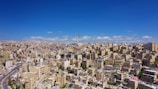 A sprawling urban landscape filled with densely packed buildings stretches out under a clear blue sky. A prominent flagpole rises above the city, displaying a colorful flag. The architecture is primarily boxy and beige, typical of Middle Eastern cities.