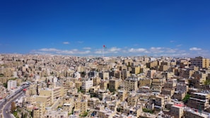 A sprawling urban landscape filled with densely packed buildings stretches out under a clear blue sky. A prominent flagpole rises above the city, displaying a colorful flag. The architecture is primarily boxy and beige, typical of Middle Eastern cities.