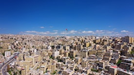 A sprawling urban landscape filled with densely packed buildings stretches out under a clear blue sky. A prominent flagpole rises above the city, displaying a colorful flag. The architecture is primarily boxy and beige, typical of Middle Eastern cities.