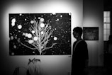 A black and white photograph depicting a man walking past large artworks in a gallery. The main artwork features a stylized tree with branches against a backdrop reminiscent of a starry night. Circular patterns are scattered across the canvas, evoking a sense of snowfall or a celestial scene. Nearby, a smaller artwork shares a night sky theme.