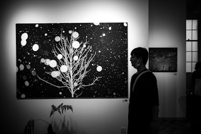 A black and white photograph depicting a man walking past large artworks in a gallery. The main artwork features a stylized tree with branches against a backdrop reminiscent of a starry night. Circular patterns are scattered across the canvas, evoking a sense of snowfall or a celestial scene. Nearby, a smaller artwork shares a night sky theme.