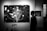 A black and white photograph depicting a man walking past large artworks in a gallery. The main artwork features a stylized tree with branches against a backdrop reminiscent of a starry night. Circular patterns are scattered across the canvas, evoking a sense of snowfall or a celestial scene. Nearby, a smaller artwork shares a night sky theme.
