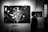 A black and white photograph depicting a man walking past large artworks in a gallery. The main artwork features a stylized tree with branches against a backdrop reminiscent of a starry night. Circular patterns are scattered across the canvas, evoking a sense of snowfall or a celestial scene. Nearby, a smaller artwork shares a night sky theme.