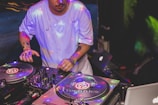 A DJ is actively mixing music on two turntables in a low-light setting, with colorful lights reflecting off the equipment. The DJ is wearing a light blue shirt and has visible tattoos on the arms.