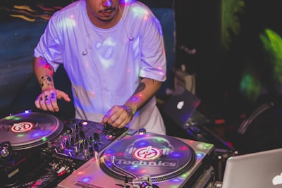 A DJ is actively mixing music on two turntables in a low-light setting, with colorful lights reflecting off the equipment. The DJ is wearing a light blue shirt and has visible tattoos on the arms.