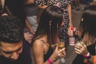A group of people are gathered together, likely at a social event or party. They are holding drinks and wearing wristbands, suggesting an informal and lively atmosphere. The setting appears to be indoors with dim lighting.
