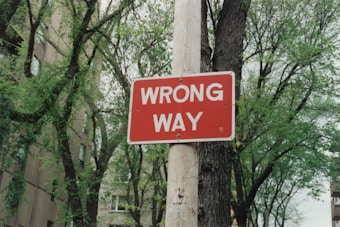 A red rectangular traffic sign is mounted on a wooden pole, displaying the words 'WRONG WAY' in bold white letters. The background features green leafy trees and a building with a brick facade.