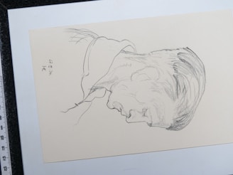 A pencil sketch depicting the profile of a man's head facing downward. The sketch is drawn on an off-white paper and includes subtle shading to indicate hair and facial features. There are markings on the left side that appear to be signatures or notes.