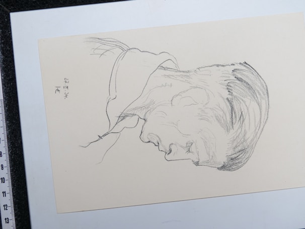 A pencil sketch depicting the profile of a man's head facing downward. The sketch is drawn on an off-white paper and includes subtle shading to indicate hair and facial features. There are markings on the left side that appear to be signatures or notes.