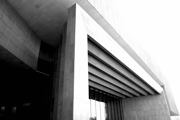 A modern architectural structure featuring geometric patterns with large, overhanging horizontal beams. The building has a sleek, minimalist design, and the perspective emphasizes its grandeur and scale. The use of concrete and glass creates a stark contrast between the light and shadow.