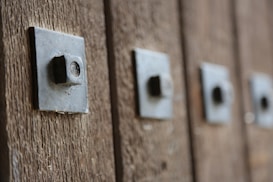 A close-up view of a series of metal bolts or studs affixed to a wooden surface, creating a textured pattern. Each bolt is embedded in a square metal plate, and the image captures a sense of rustic or industrial design.