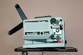 A vintage film projector is placed on a dark surface. It features a metal casing with various knobs, reels, and switches. The text 'Heurtier' and 'Super8' is visible on the body, indicating the brand and format. The arms for the film reels extend outwards.