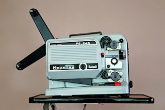 A vintage film projector is placed on a dark surface. It features a metal casing with various knobs, reels, and switches. The text 'Heurtier' and 'Super8' is visible on the body, indicating the brand and format. The arms for the film reels extend outwards.