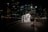A nighttime urban scene with a brightly lit wooden shelter on a boardwalk against the backdrop of tall city buildings illuminated by numerous lights. The shelter has a no smoking sign prominently displayed. A bench is positioned nearby, and streetlights cast a warm glow on the pathway.