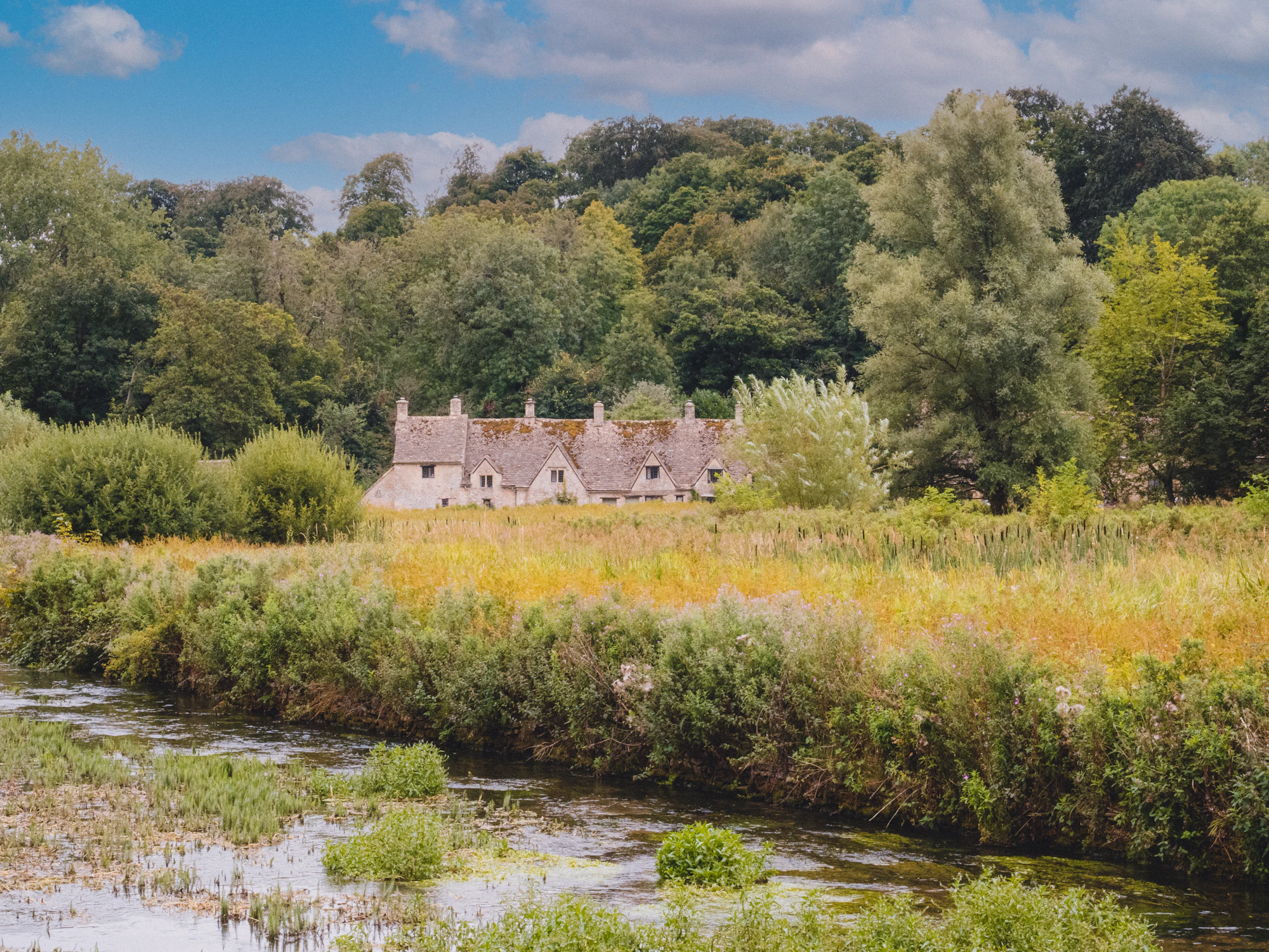 A honey-stone house set across a summer water meadow and slow river in the Cotswolds