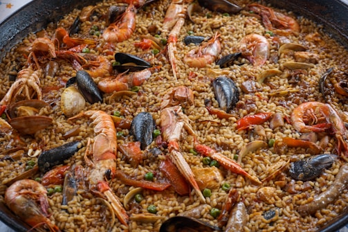 A close-up view of a large pan filled with a seafood paella. The dish contains a mixture of shrimp, mussels, clams, and rice cooked with a rich blend of spices. The seafood is arranged on top of the rice, and small pieces of vegetables, such as peas, are scattered throughout.