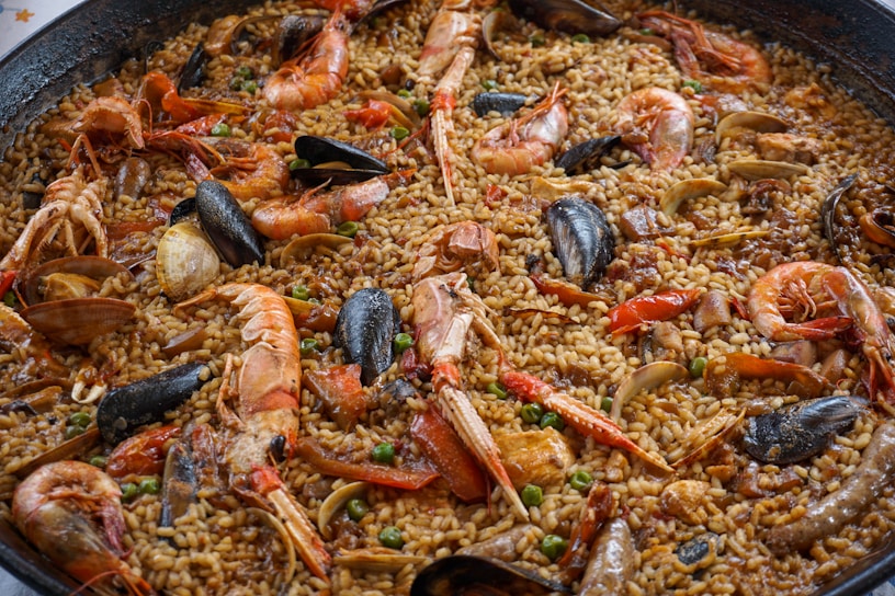 A close-up view of a large pan filled with a seafood paella. The dish contains a mixture of shrimp, mussels, clams, and rice cooked with a rich blend of spices. The seafood is arranged on top of the rice, and small pieces of vegetables, such as peas, are scattered throughout.