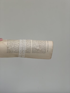 A page from a book is held up against a plain gray background. The page features columns of typed text with a decorative white lace wrapped around it. The hand holding the page is positioned to the left, and the page appears slightly curved.