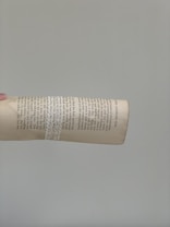 A page from a book is held up against a plain gray background. The page features columns of typed text with a decorative white lace wrapped around it. The hand holding the page is positioned to the left, and the page appears slightly curved.