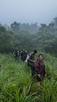 A group of people is hiking through tall grass in a lush, misty forest. They are wearing raincoats and appear to be enjoying their trek in the natural surroundings. The background is filled with dense trees under a cloudy sky, creating a serene and adventurous atmosphere.