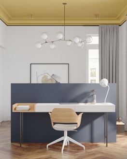 A modern and minimalist home office setup features a stylish wooden chair with a white cushioned seat, in front of a sleek white desk. The desk is equipped with a white desk lamp, a closed laptop, a small vase with dried plants, and two stacked books. The background wall is painted in a deep blue color and a piece of abstract art hangs on it. The room is well-lit, with a distinctive hanging chandelier consisting of several round glass bulbs, and a striking yellow ceiling. The floor is made of a warm-toned herringbone wood pattern.