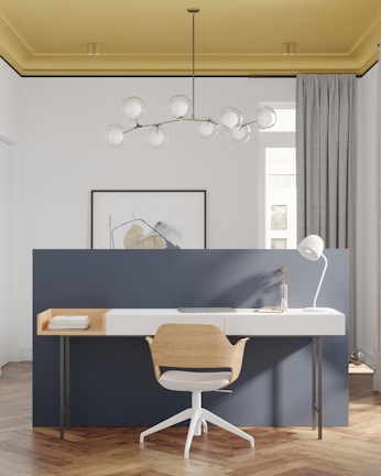 A modern and minimalist home office setup features a stylish wooden chair with a white cushioned seat, in front of a sleek white desk. The desk is equipped with a white desk lamp, a closed laptop, a small vase with dried plants, and two stacked books. The background wall is painted in a deep blue color and a piece of abstract art hangs on it. The room is well-lit, with a distinctive hanging chandelier consisting of several round glass bulbs, and a striking yellow ceiling. The floor is made of a warm-toned herringbone wood pattern.