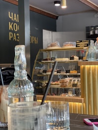 A cozy cafe interior with a modern design. A glass display case is filled with various pastries and desserts, each labeled with prices in a foreign currency. In the foreground, there are empty glasses and utensils on a marble table. A decorative glass bottle and a pink phone are also visible. Soft, warm lighting creates a welcoming atmosphere.
