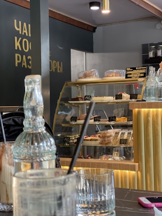 A cozy cafe interior with a modern design. A glass display case is filled with various pastries and desserts, each labeled with prices in a foreign currency. In the foreground, there are empty glasses and utensils on a marble table. A decorative glass bottle and a pink phone are also visible. Soft, warm lighting creates a welcoming atmosphere.