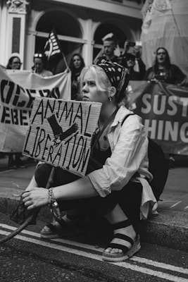 A person with light hair is kneeling on a street holding a sign that reads 'Animal Liberation'. The person is surrounded by other individuals, some holding flags and banners with messages. The atmosphere suggests a protest or demonstration taking place.