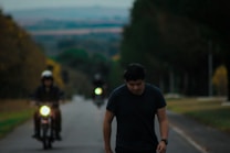 A person walks along a tree-lined road, appearing deep in thought. In the background, two motorcycles are approaching with their headlights on. The scene has a moody ambiance, with overcast skies and lush green and brown foliage framing the narrow road.