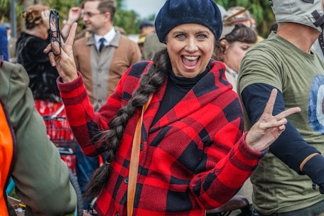 A person with a long braid and a black beret smiles broadly while holding up a peace sign with both hands. They are wearing a red and black checkered coat and stand among a group of people, some of whom are dressed in vintage-style clothing.