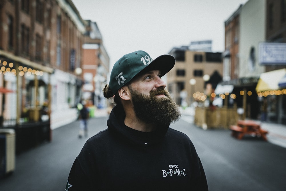 man in black crew neck shirt wearing green and red cap standing on road during daytime