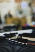 A close-up of a straight razor with a wooden handle lying on a textured black surface. Surrounding the razor are various grooming tools, including brushes. The background is blurred, creating a bokeh effect with circular light spots.