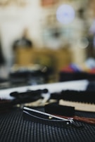 A close-up of a straight razor with a wooden handle lying on a textured black surface. Surrounding the razor are various grooming tools, including brushes. The background is blurred, creating a bokeh effect with circular light spots.