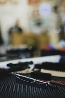 A close-up of a straight razor with a wooden handle lying on a textured black surface. Surrounding the razor are various grooming tools, including brushes. The background is blurred, creating a bokeh effect with circular light spots.