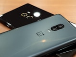 Two smartphones placed on a wooden surface, one is grey metallic with dual rear cameras and a brand logo, while the second is glossy black with a single camera and circular design around it.
